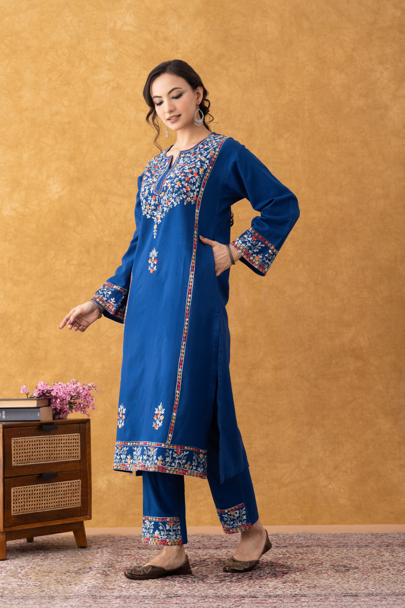 Woman wearing a blue embroidered traditional outfit against a beige wall. Blue embroidered kurta set, royal blue kurta set with dupatta, navy blue kurta set with dupatta, sky blue kurta set with dupatta, winter suits for women, winter suits designs for ladies, winter party wear suits for ladies, winter stitched suits for ladies, winter wear suits for women, Kashmiri winter suits for ladies.